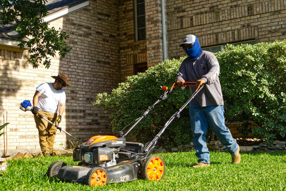 Two lawn crew members mowing and trimming a front lawn beside a brick house
